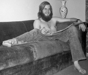 Bearded white male with long hair and glasses, shirtless and barefoot, in jeans sitting on a couch with the newspaper. He is wearing his late grandmother's wedding ring on a chain around his neck and smiling.